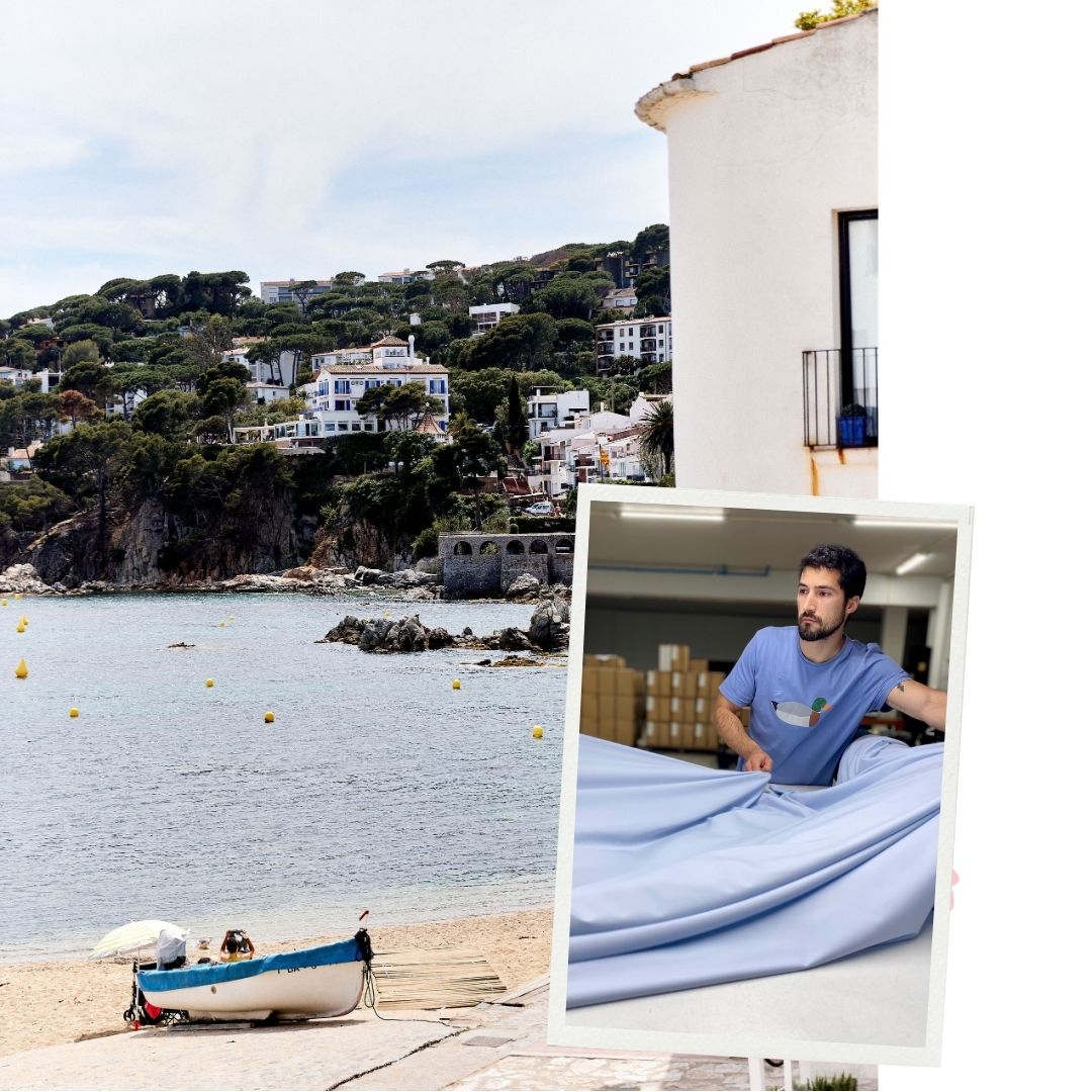 Un paysage côtier avec une plage, une mer calme, présence de bouées jaunes, et des maisons blanches et bleues entourées de verdure sur une colline en arrière-plan. Un petit bateau de pêche sur le sable à côté d'un parasol. Sur la droite, une maison blanche avec une fenêtre et un pot de fleurs bleues. Superposé à l'image, un cadre photo incliné montre un homme barbu portant un t-shirt bleu avec un logo coloré, en train de manipuler un grand tissu bleu clair dans un atelier rempli de cartons.