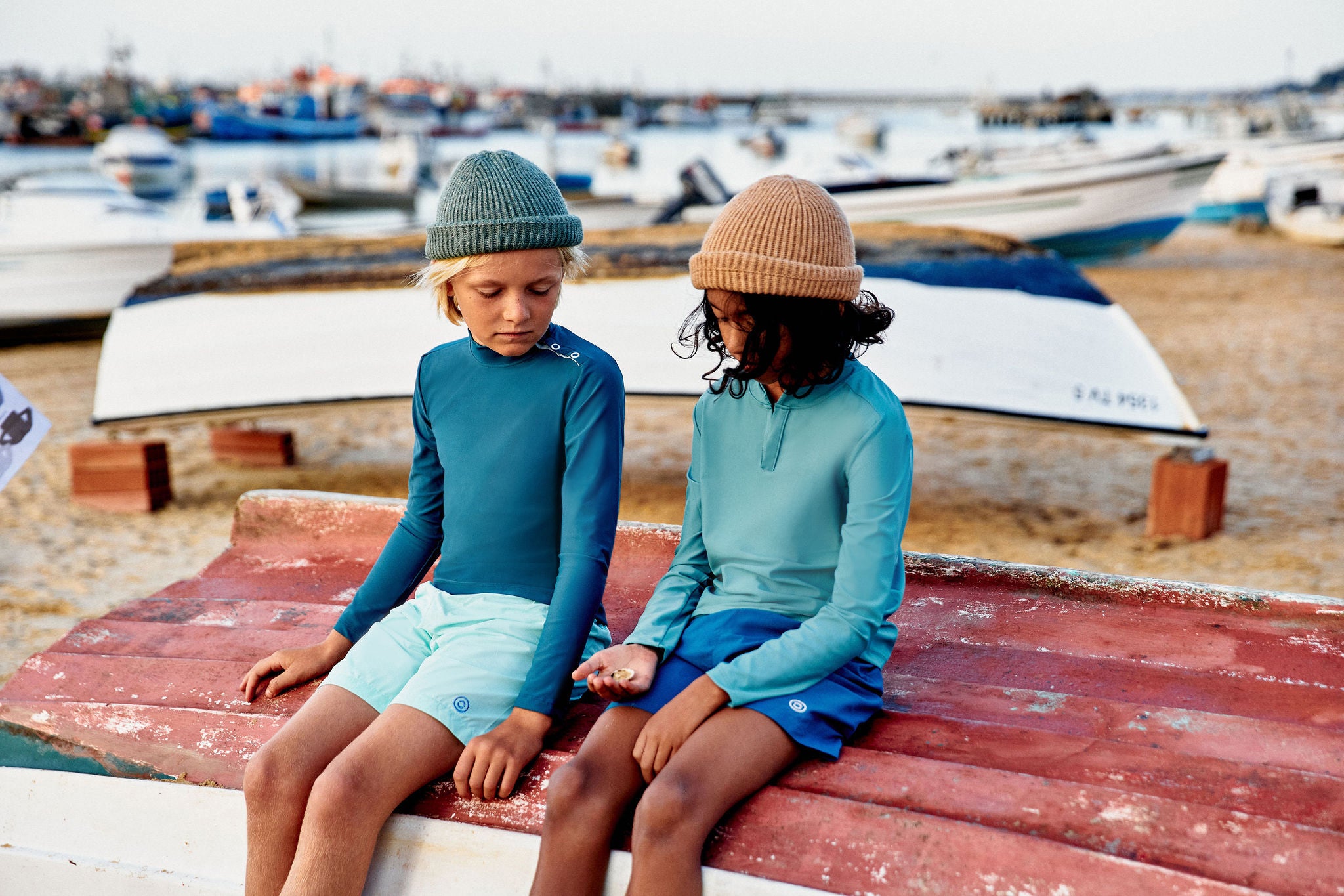 Deux garçons assis sur une barque rouge sur le sable, dans un port où de nombreux bateaux sont en arrière-plan. Ils portent des tenues de protection solaire. Tous deux portent des bonnets, leur donnant un air à la fois décontracté et aventureux. L’enfant à droite observe  un petit objet dans sa main, et l’autre regarde avec curiosité. 