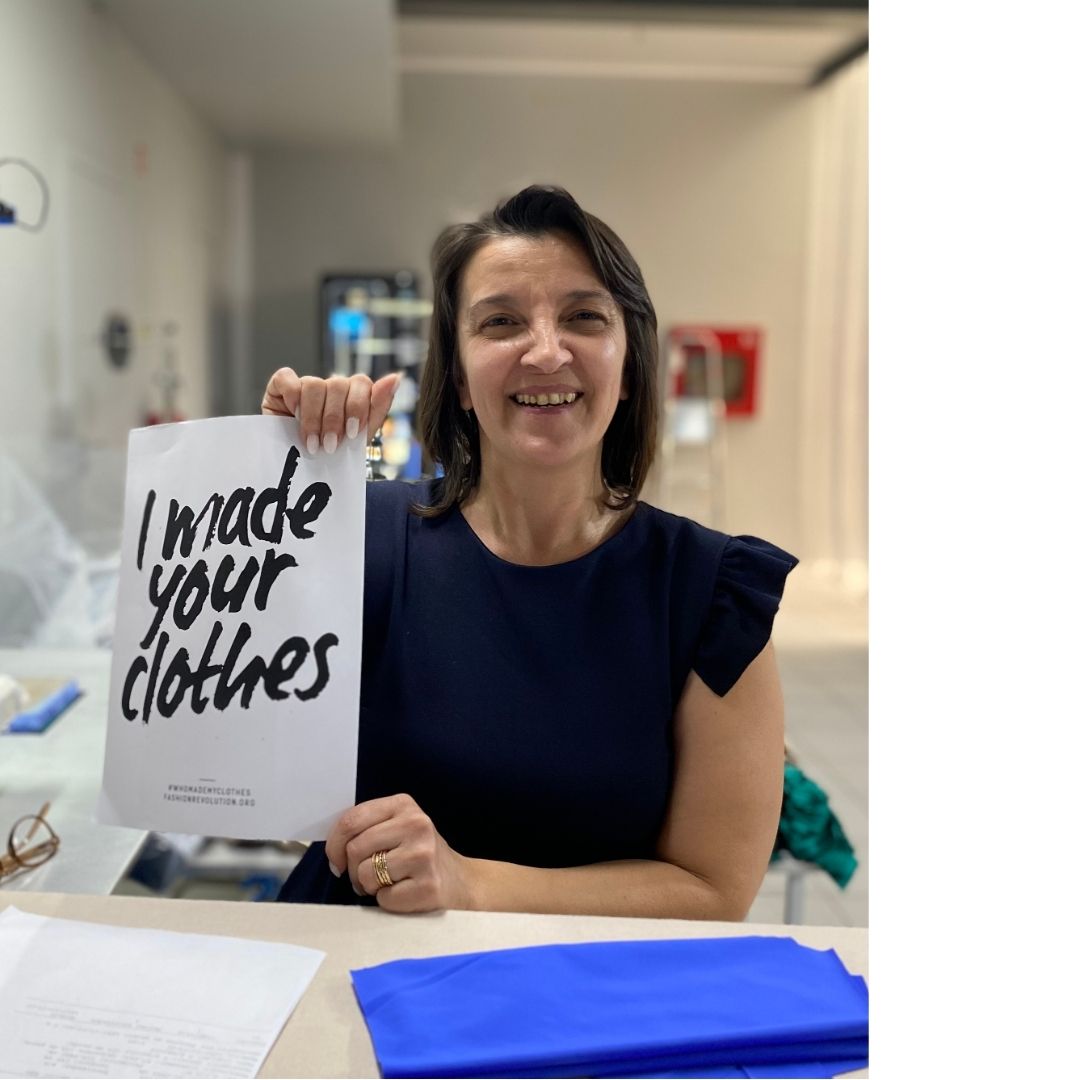 Une femme souriante, aux cheveux bruns, porte une robe bleu marine et est assise à une table dans un atelier de confection. Elle tient une feuille de papier blanche avec l'inscription en lettres noires : 'I made your clothes' (J’ai fabriqué vos vêtements). En arrière-plan, on aperçoit du matériel de couture et des rouleaux de tissu. La scène met en avant le travail artisanal et l'engagement pour une mode plus transparente et éthique.