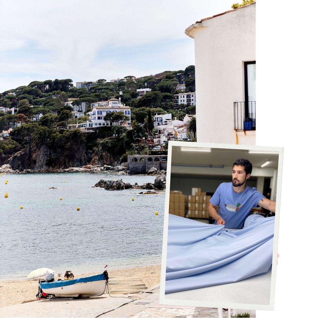 Un paysage côtier avec une plage, une mer calme, présence de bouées jaunes, et des maisons blanches et bleues entourées de verdure sur une colline en arrière-plan. Un petit bateau de pêche  sur le sable à côté d'un parasol. Sur la droite, une maison blanche avec une fenêtre et un pot de fleurs bleues. Superposé à l'image, un cadre photo incliné montre un homme barbu portant un t-shirt bleu avec un logo coloré, en train de manipuler un grand tissu bleu clair dans un atelier rempli de cartons.