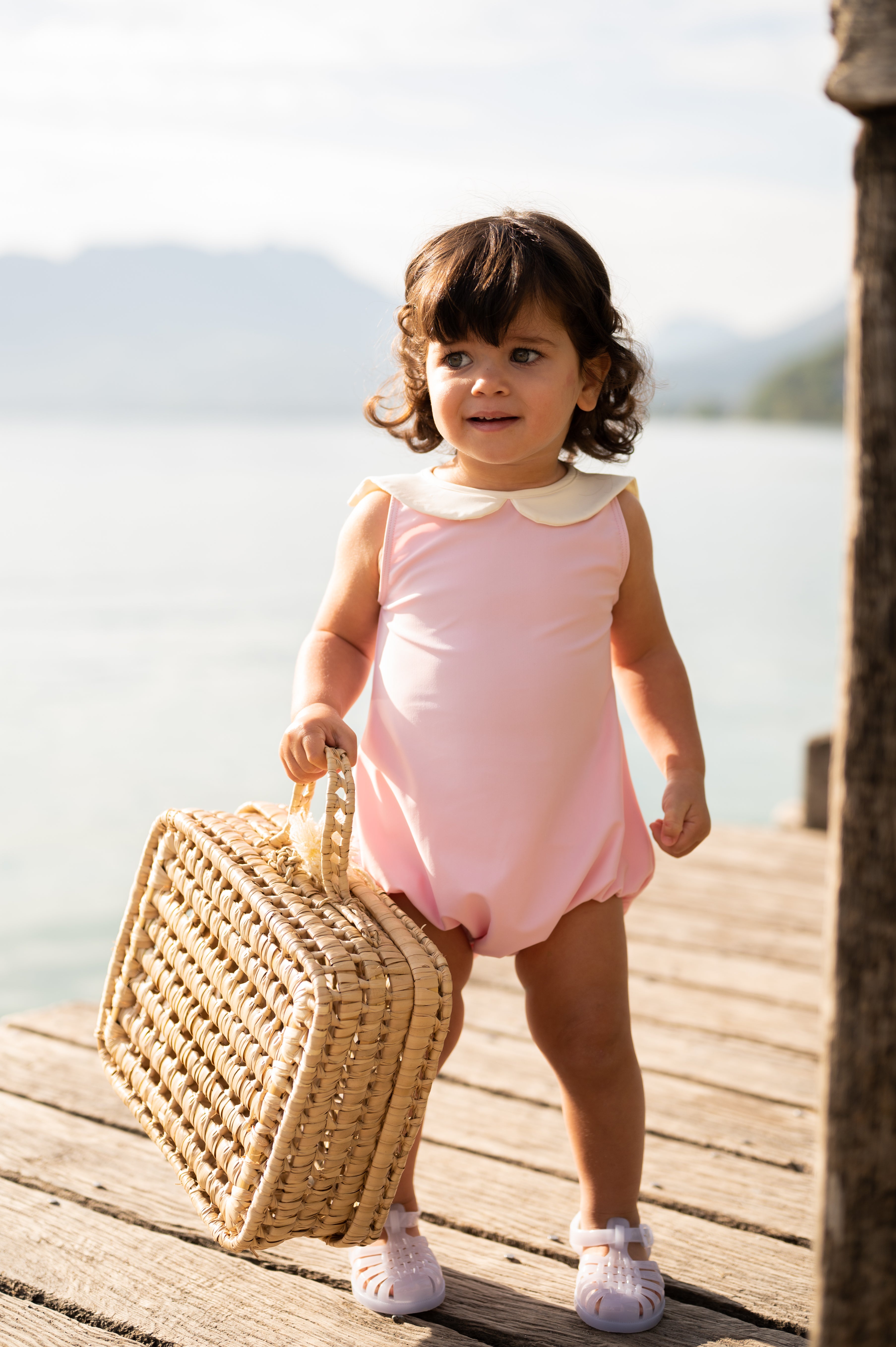 Une petite fille aux cheveux bouclés bruns et aux yeux pétillants marche sur un ponton en bois au bord d’un lac, tenant un sac dans sa main. Elle porte un maillot de bain rose à col blanc nommé simone et des sandales transparentes. En arrière-plan, le lac scintille sous le soleil, avec des montagnes floues à l’horizon. La scène dégage une ambiance estivale et insouciante.